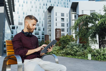 Geschäftsmann mit Kopfhörern, der ein digitales Tablet benutzt, während er auf einer Bank in der Stadt sitzt - BOYF01653