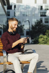 Smiling businessman using smart phone while sitting on bench during sunny day - BOYF01654