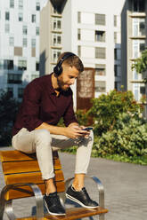 Businessman wearing headphones using smart phone while sitting on bench - BOYF01657