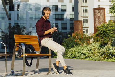 Entrepreneur wearing headphone using mobile phone while sitting on bench in city - BOYF01660