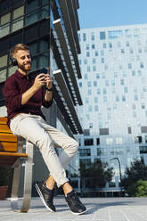 Male entrepreneur wearing headphones using mobile phone while sitting on bench in city - BOYF01666