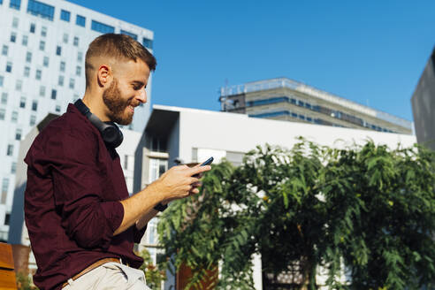 Mittlerer erwachsener Geschäftsmann mit Kopfhörern, der in der Stadt sitzend ein Smartphone benutzt - BOYF01667