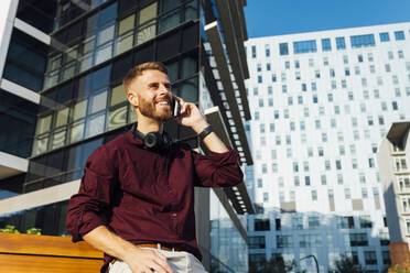 Lächelnder Geschäftsmann, der auf einer Bank sitzend mit seinem Handy telefoniert - BOYF01670