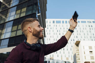Smiling businessman taking selfie through mobile phone while sitting in city - BOYF01675