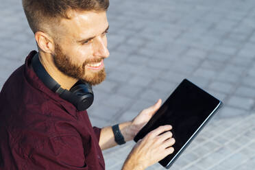 Lächelnder Geschäftsmann, der wegschaut, während er ein digitales Tablet benutzt, das auf einem Fußweg steht - BOYF01676