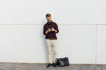 Businessman with headphones using mobile phone while standing against white wall - BOYF01680