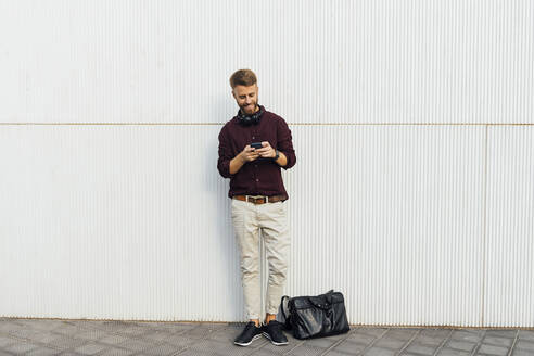 Geschäftsmann mit Kopfhörern, der ein Mobiltelefon benutzt, während er an einer weißen Wand steht - BOYF01680