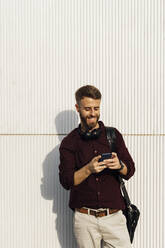Smiling businessman with headphones and shoulder bag using mobile phone while standing against white wall - BOYF01684