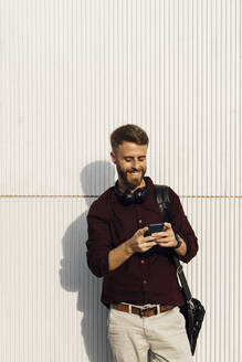 Lächelnder Geschäftsmann mit Kopfhörern und Umhängetasche, der ein Mobiltelefon benutzt, während er vor einer weißen Wand steht - BOYF01684