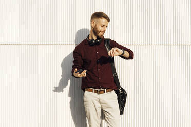 Mid adult businessman with mobile phone and shoulder bag checking time while standing against white wall - BOYF01685