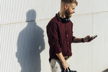 Businessman with headphones using mobile phone while standing by white wall - BOYF01688