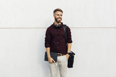 Confident businessman with digital tablet and shoulder bag looking away while standing against white wall - BOYF01689