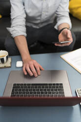 Geschäftsmann, der sein Handy in der Hand hält, während er im Büro zu Hause am Laptop arbeitet - BOYF01722