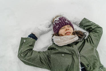 Girl in green jacket with eyes closed making snow angle during vacations - OGF00842