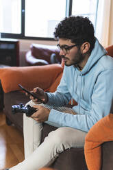 Junger Mann, der ein Smartphone mit einem Spiel-Controller benutzt, während er auf dem Sofa im Wohnzimmer sitzt - JAQF00196