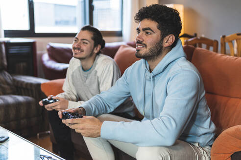 Male friends enjoying video game while sitting on sofa in living room - JAQF00204