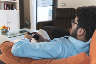 Entspannter junger Mann spielt ein Videospiel, während er auf dem Sofa im Wohnzimmer sitzt - JAQF00206