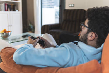 Relaxed young man playing video game while sitting on sofa in living room - JAQF00206