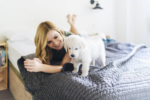 Smiling woman looking at Golden Retriever puppy while lying on bed - DGOF01897