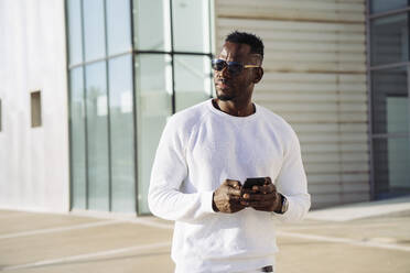 Afrikanischer Mann mit Sonnenbrille und Smartphone vor einem Gebäude - MPPF01419