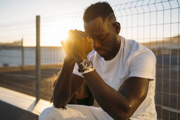 Worried African man sitting against fence during sunset - MPPF01444