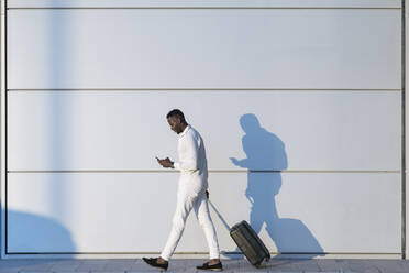 Man with suitcase using smart phone while walking by white wall - MPPF01453
