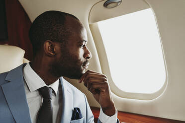 Männlicher Unternehmer mit Hand am Kinn, der durch ein Fenster im Flugzeug schaut - OIPF00238
