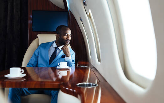 Young businessman with hand on chin looking through window while sitting in private jet - OIPF00239