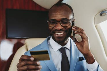 Smiling businessman holding credit card while talking on mobile phone in airplane - OIPF00271