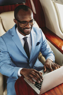 Lächelnder männlicher Unternehmer, der im Privatjet an einem Laptop arbeitet - OIPF00273
