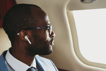 Männlicher Unternehmer mit drahtlosen Kopfhörern, der durch das Fenster eines Privatjets schaut - OIPF00278