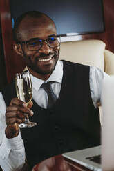Lächelnder männlicher Unternehmer mit Champagner, der im Privatjet an einem Laptop arbeitet - OIPF00320