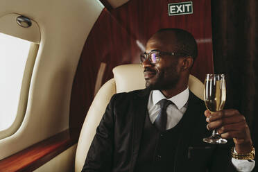 Lächelnder männlicher Unternehmer mit Champagner in der Hand, der durch das Fenster eines Privatjets schaut - OIPF00334