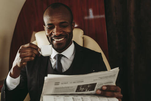 Glücklicher männlicher Unternehmer mit Kaffeetasse beim Lesen der Zeitung im Privatjet - OIPF00339