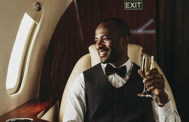 Lächelnder männlicher Unternehmer mit Champagner in der Hand, der aus dem Fenster eines Flugzeugs schaut - OIPF00342