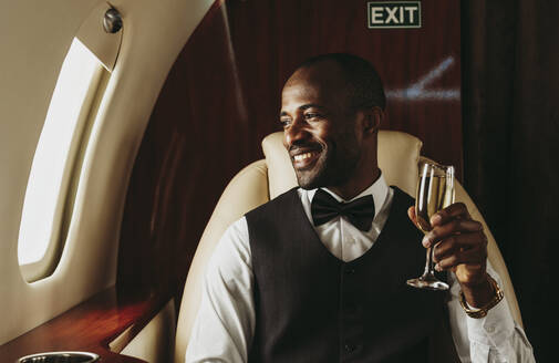 Lächelnder männlicher Unternehmer mit Champagner in der Hand, der aus dem Fenster eines Flugzeugs schaut - OIPF00342