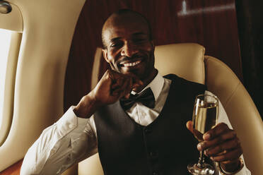 Männlicher Unternehmer mit Hand am Kinn hält Champagner im Flugzeug - OIPF00344