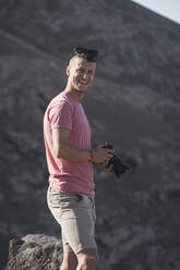 Happy male tourist holding camera at Femes Viewpoint on sunny day, Lanzarote, Spain - SNF01077