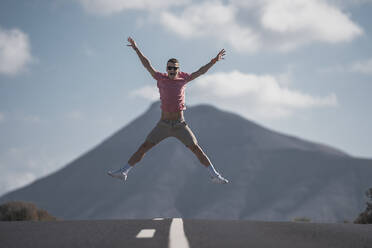 Fröhlicher junger männlicher Tourist, der an einem sonnigen Tag auf Lanzarote über die Straße gegen vulkanisches Gestein springt, Spanien - SNF01079