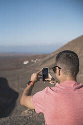 Young male tourist photographing through smart phone at Femés viewpoint, Lanzarote on sunny day, Spain - SNF01087