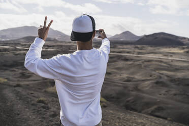 Young male tourist peace gesturing while taking selfie at El Cuervo Volcano during vacations, Lanzarote, Spain - SNF01093