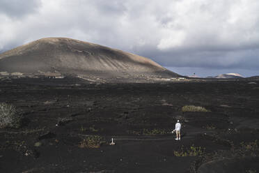 Young male tourist standing on black soil in vineyard at El Cuervo Volcano during vacations, Lanzarote, Spain - SNF01102