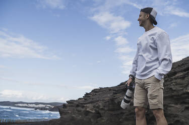 Lächelnder männlicher Tourist mit Händen in den Taschen, der eine Kamera hält, während er die Aussicht in El golfo, Lanzarote, Spanien, betrachtet - SNF01110