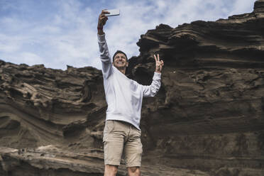 Smiling male tourist gesturing peace while taking selfie against rock at El golfo, Lanzarote, Spain - SNF01119