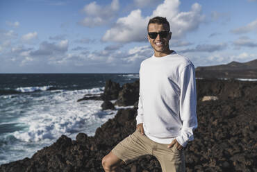 Lächelnder männlicher Tourist mit Händen in den Taschen auf einem Aussichtspunkt am Meer in Los Hervideros, Lanzarote, Spanien - SNF01120