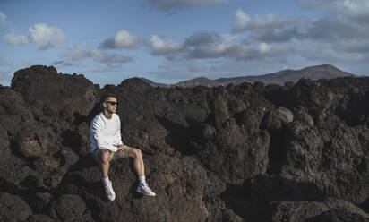 Junger männlicher Tourist mit Sonnenbrille, der die Aussicht betrachtet, während er auf einem Felsen bei Los Hervideros an einem sonnigen Tag sitzt, Lanzarote, Spanien - SNF01122