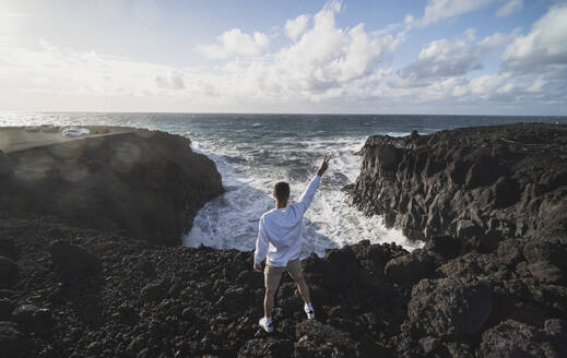 Junger männlicher Tourist, der auf einem Felsen vor dem Meer in Los Hervideros, Lanzarote, Spanien, steht und Frieden gestikuliert - SNF01126