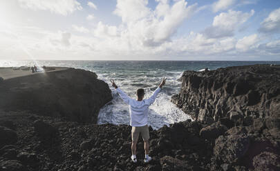 Male tourist making peace sign while standing on volcanic rock during sunny day at Los Hervideros, Lanzarote, Spain - SNF01127