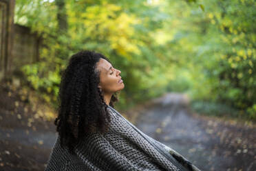 Frau mit lockigem Haar und Jacke sitzt mit geschlossenen Augen auf einem Fußweg im Wald - AKLF00003