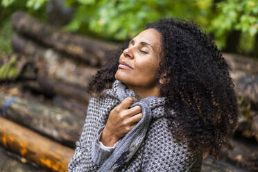 Curly hair woman smiling while sitting with eyes closed in forest - AKLF00005